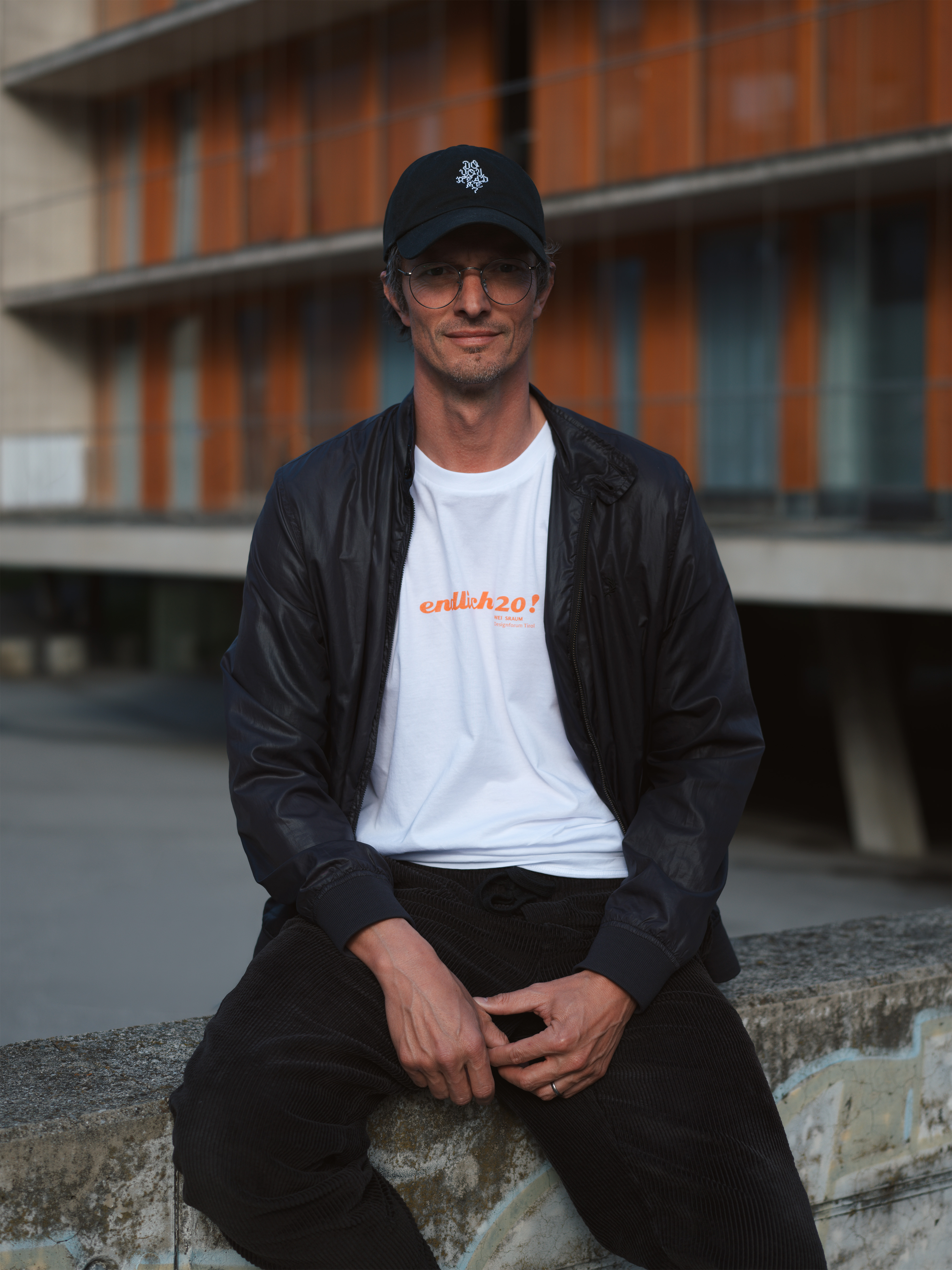Portr&auml;t Alex Kofler auf einem Stein sitzend, schwarzes Basecap, weisses T-Shirt, schwarze Jacke